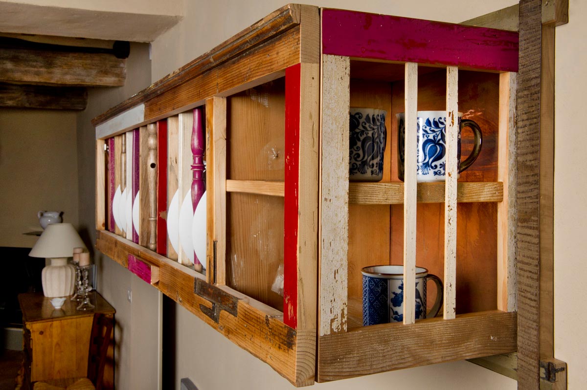 Dettaglio laterale di una piattaia in legno antico naturale e colorato, fucsia, bianco, rosa e bordeaux, con schiena con pannello di rame lucido